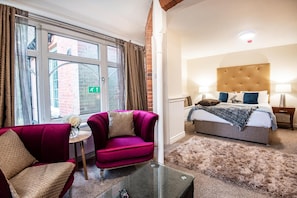 A hotel room with a bed, a sitting area with a chair and a small table, and a large window with curtains. - Trivelles Waterhall Country Hotel (Crawley)