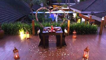 Un área de comedor al aire libre con un arco en forma de corazón y mesas dispuestas para una comida, rodeada de faroles y follaje.