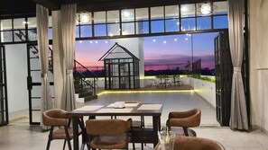 An indoor dining area with a table and chairs, large windows, and a view of a sunset. - Dawala Hotel (Makassar)