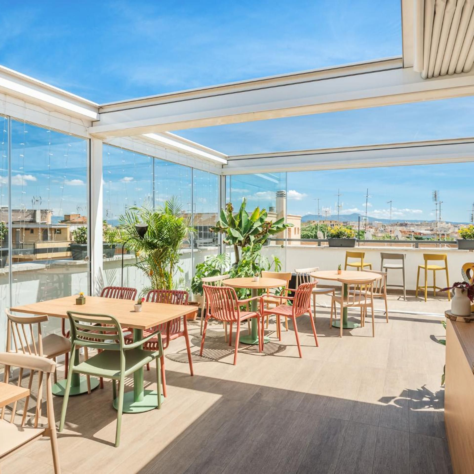 A modern outdoor dining area with tables and chairs, surrounded by glass walls and potted plants.