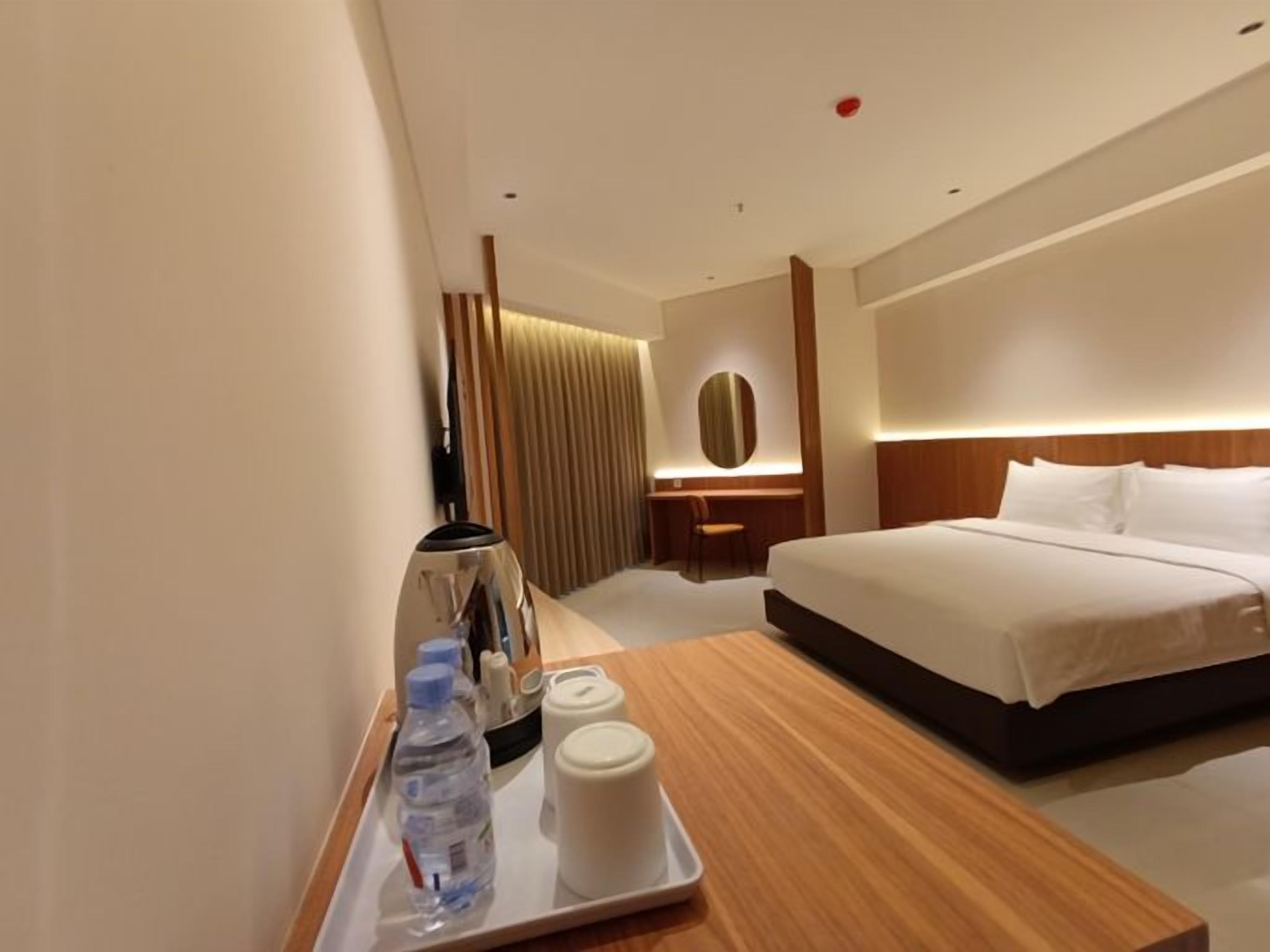 A hotel room with a bed, a bedside table with a tray of toiletries, a desk, and a mirror.