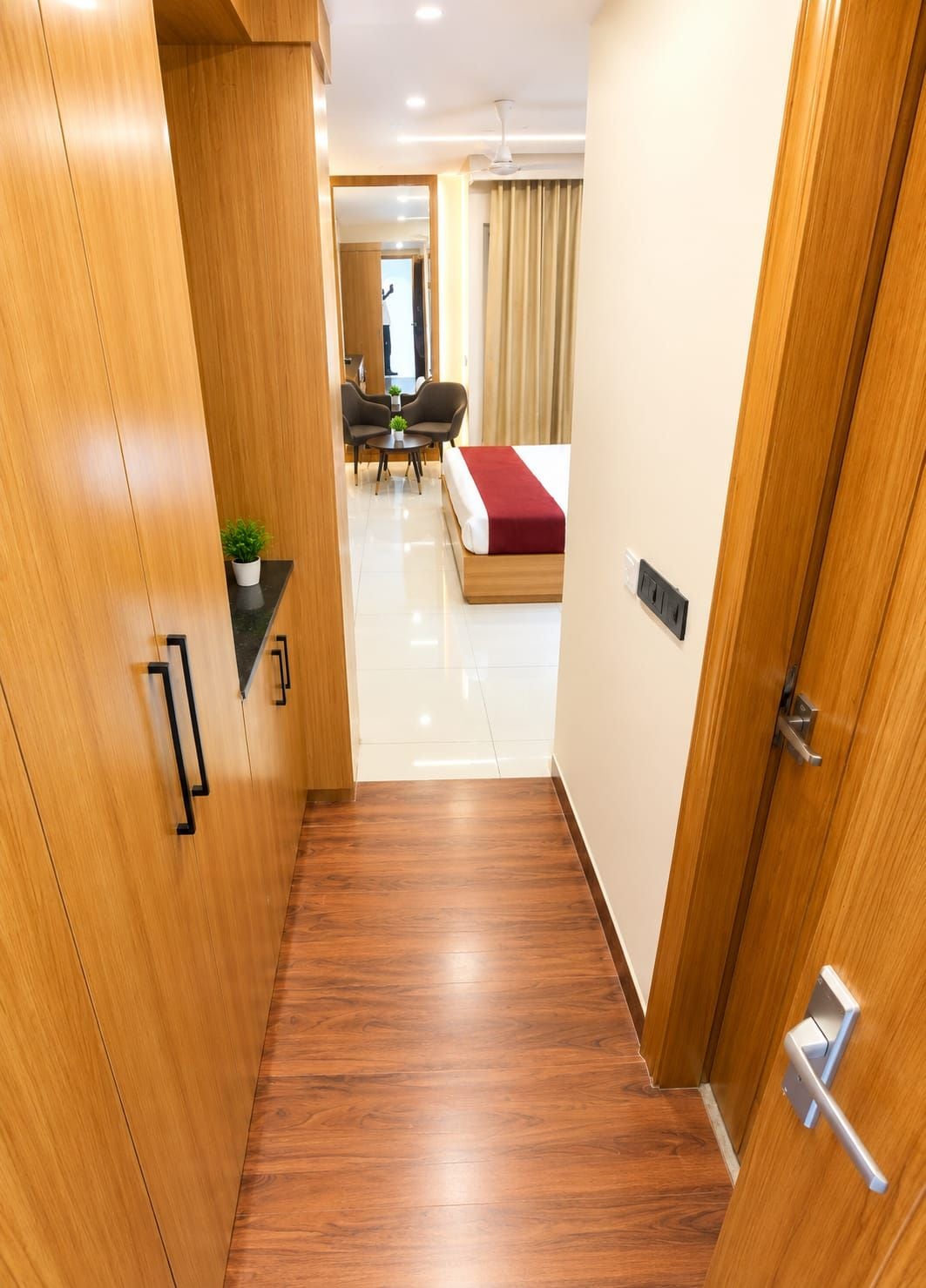 Un pasillo de hotel con piso de madera, una cama con una colcha roja, un área de comedor con sillas y una mesita con una planta.