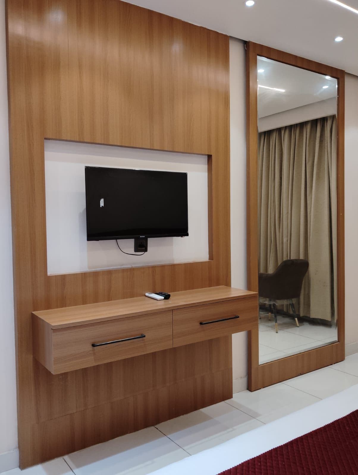 A modern room with a flat-screen TV mounted on a wooden wall, a wooden console table with drawers, and a large mirror reflecting a chair and curtains.