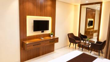 Habitación de hotel con cama, televisor montado en un panel de madera, escritorio con silla, mesita con planta y espejo.