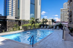 A swimming pool with a lifeguard chair, surrounded by a fence and palm trees, with tall buildings in the background. - Luxury Waikiki 1BR: Ocean View & Free Parking (Honolulu)
