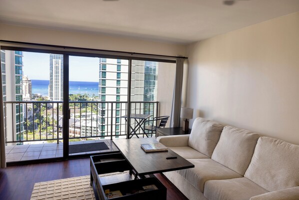 A modern living room with a sofa, a coffee table, and a view of the ocean through large windows. - Luxury Waikiki 1BR: Ocean View & Free Parking (Honolulu)