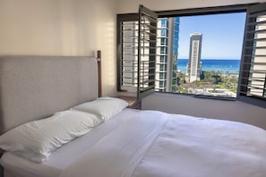 A hotel room with a bed, a window with blinds, and a view of the ocean and buildings. - Luxury Waikiki 1BR: Ocean View & Free Parking (Honolulu)