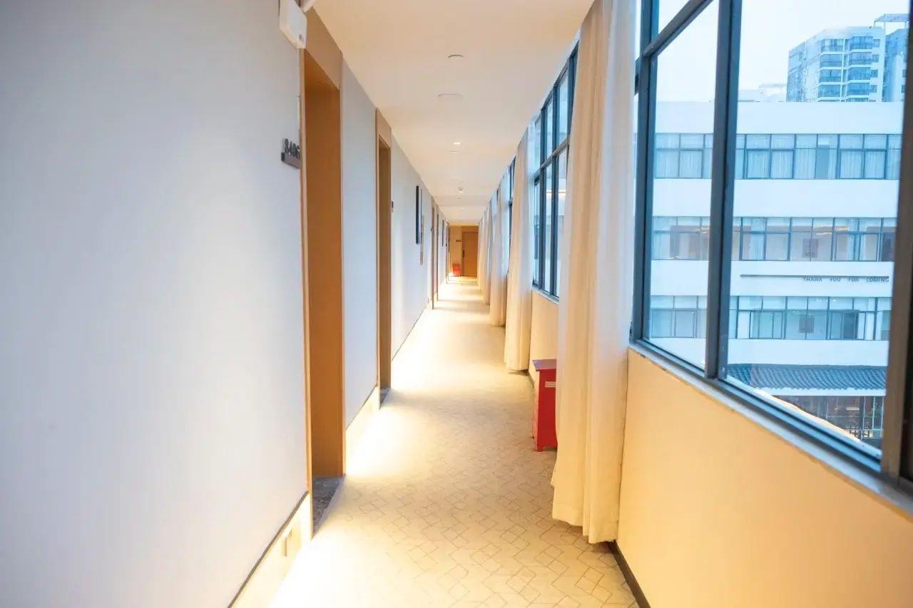 Pasillo de hotel con suelo de baldosas, ventanas grandes y un cubo de basura rojo.