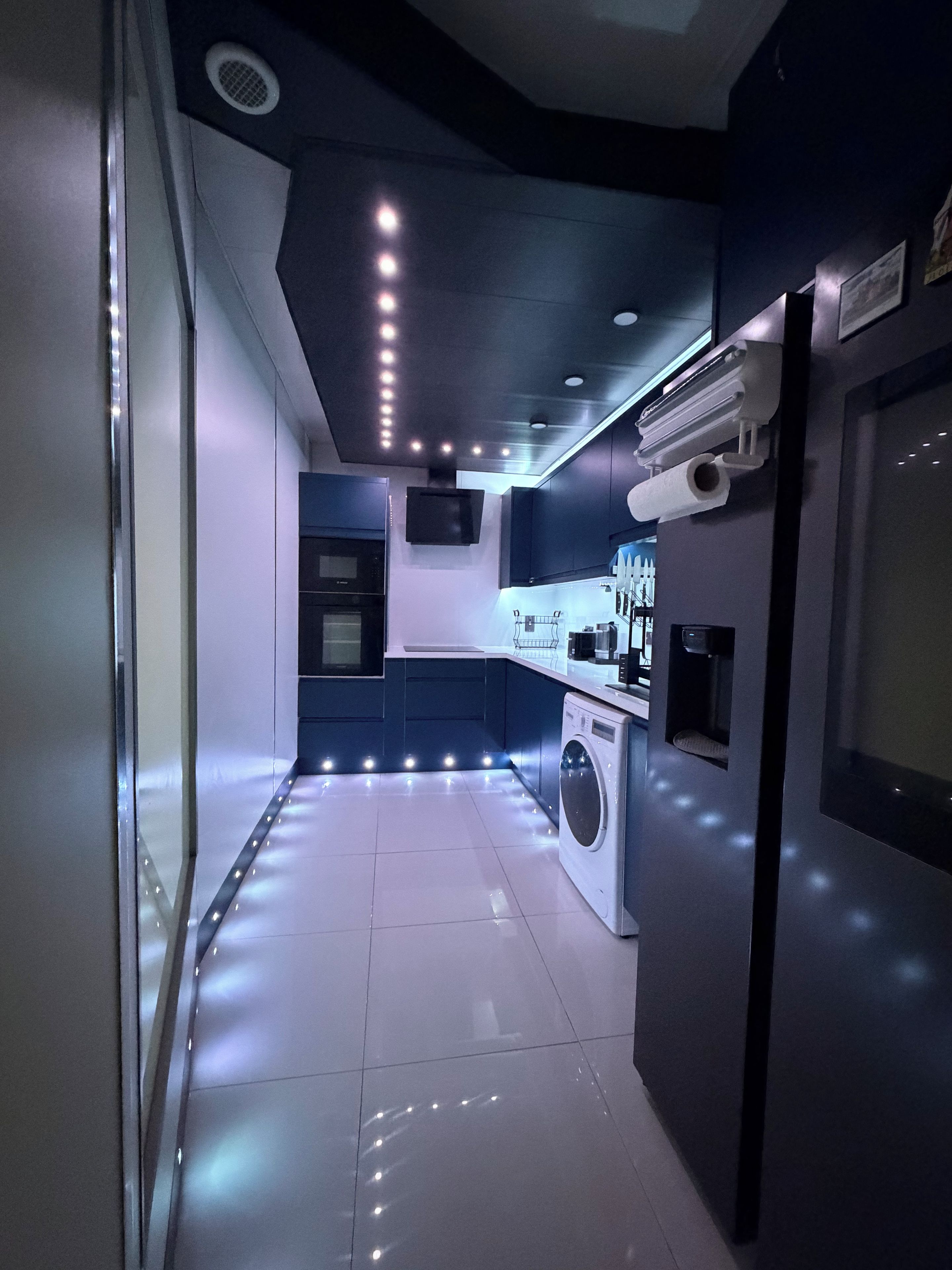 A modern kitchen with a built-in washer and dryer, recessed lighting, and a flat-screen TV mounted on the wall.
