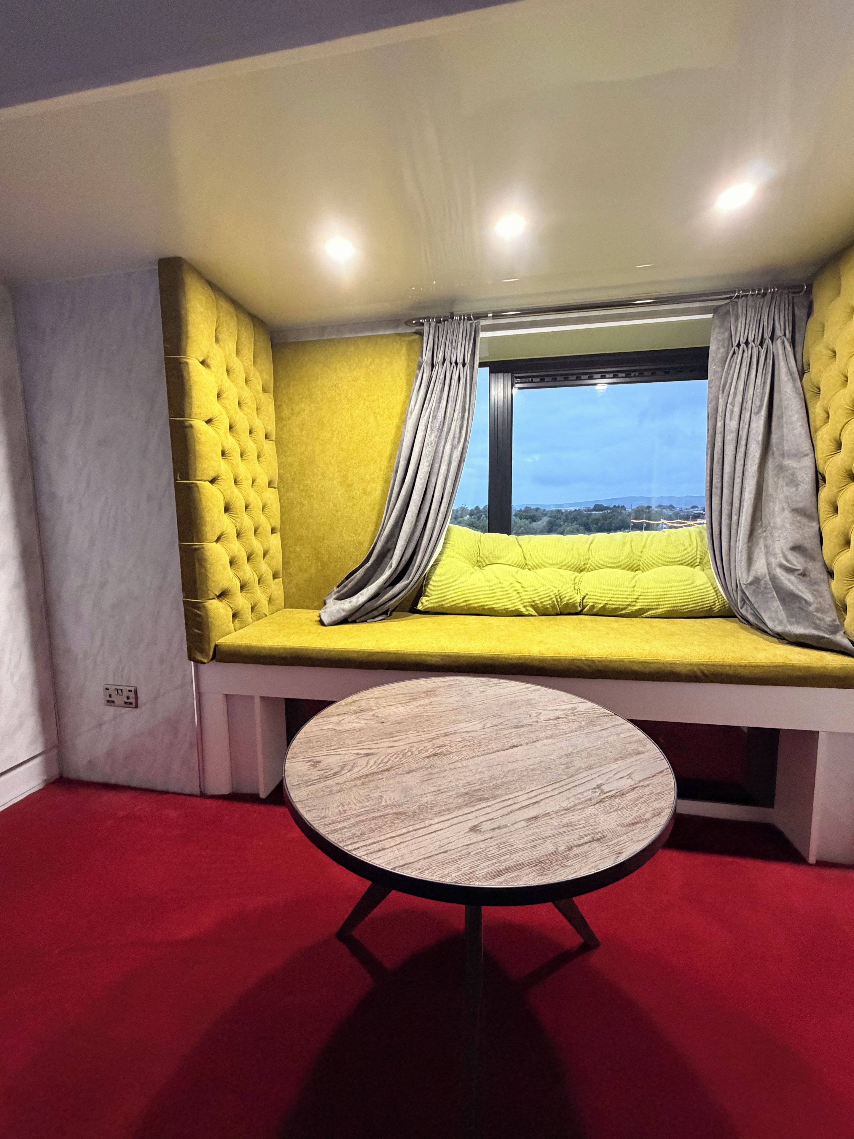 Een kamer met een geel gestoffeerde bank, een ronde tafel en een raam met gordijnen.