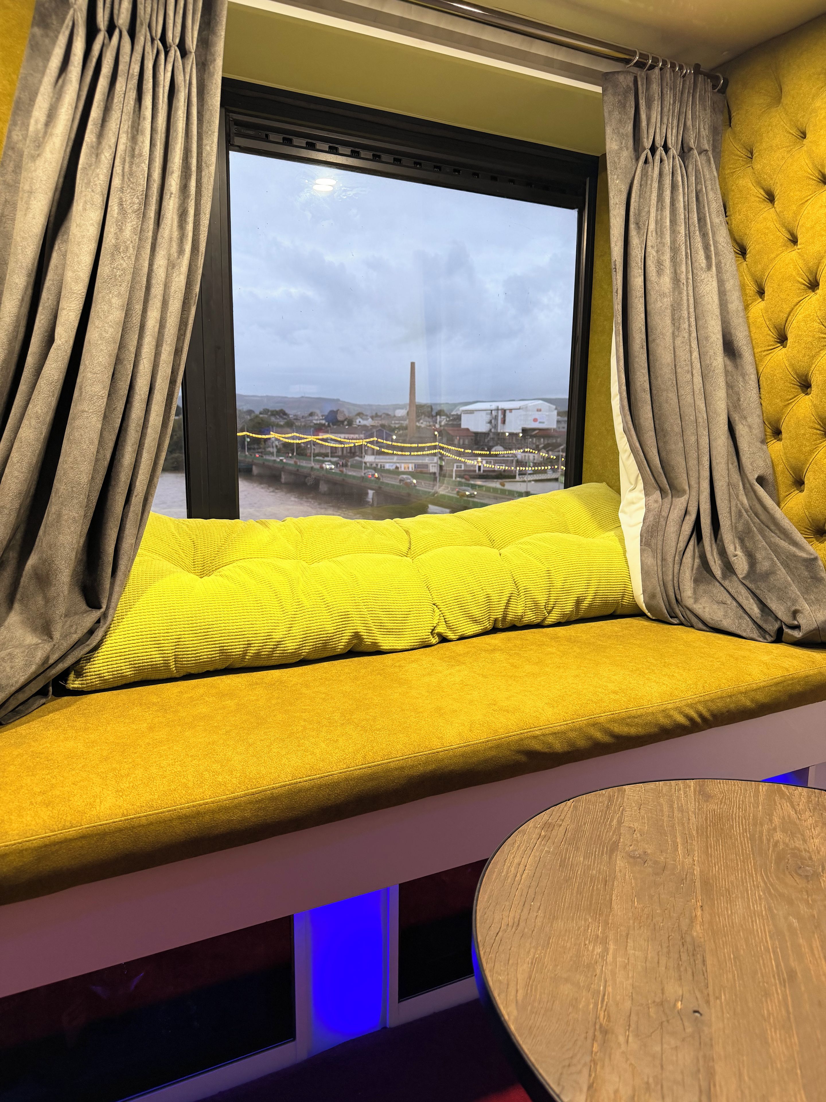 A window seat with curtains, a yellow cushion, and a view of a bridge and industrial buildings.