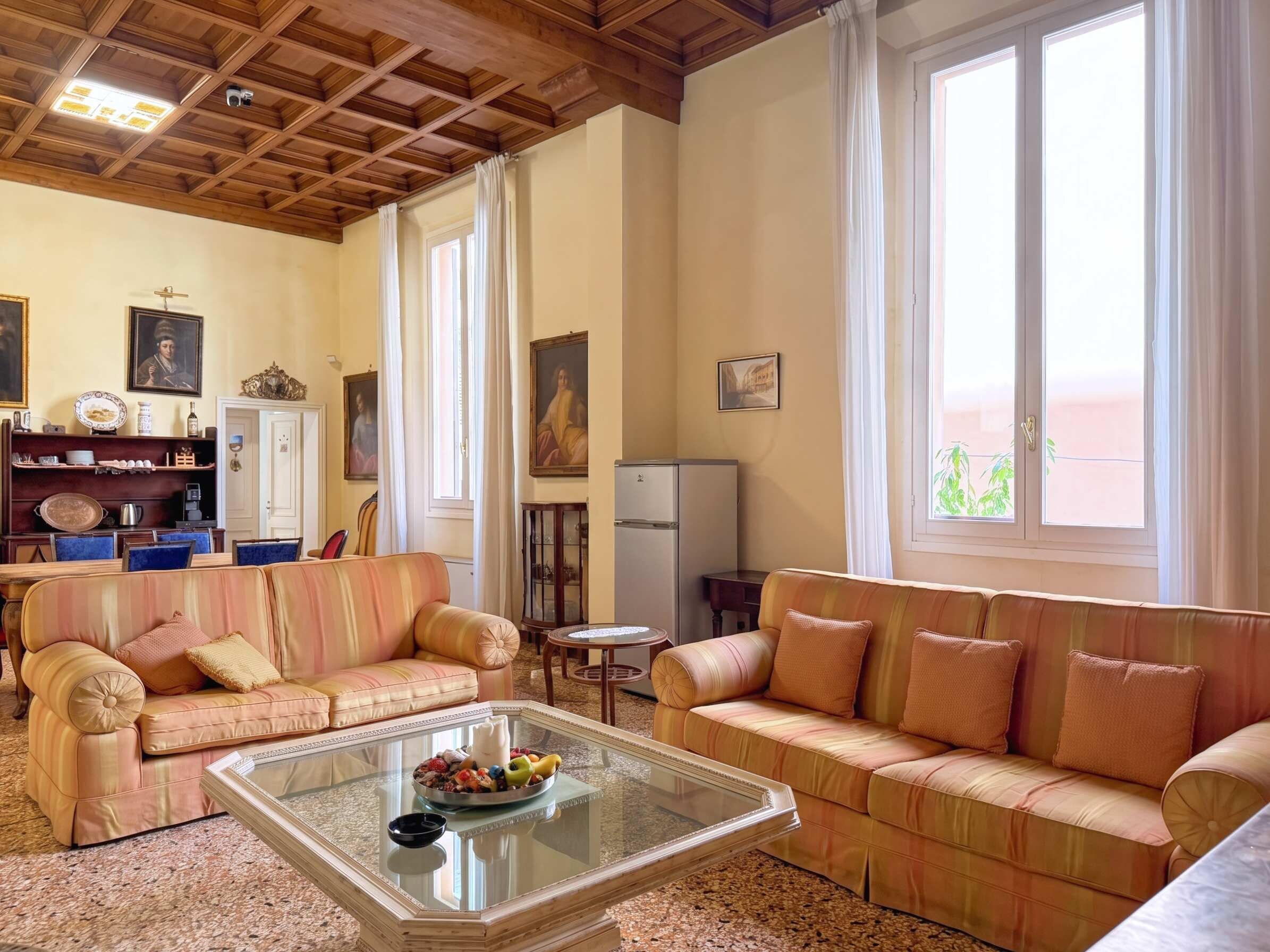 Ein Wohnzimmer mit zwei Sofas, einem Couchtisch und einem Esstisch. An den Wänden befinden sich Gemälde und im Hintergrund steht ein Kühlschrank.
