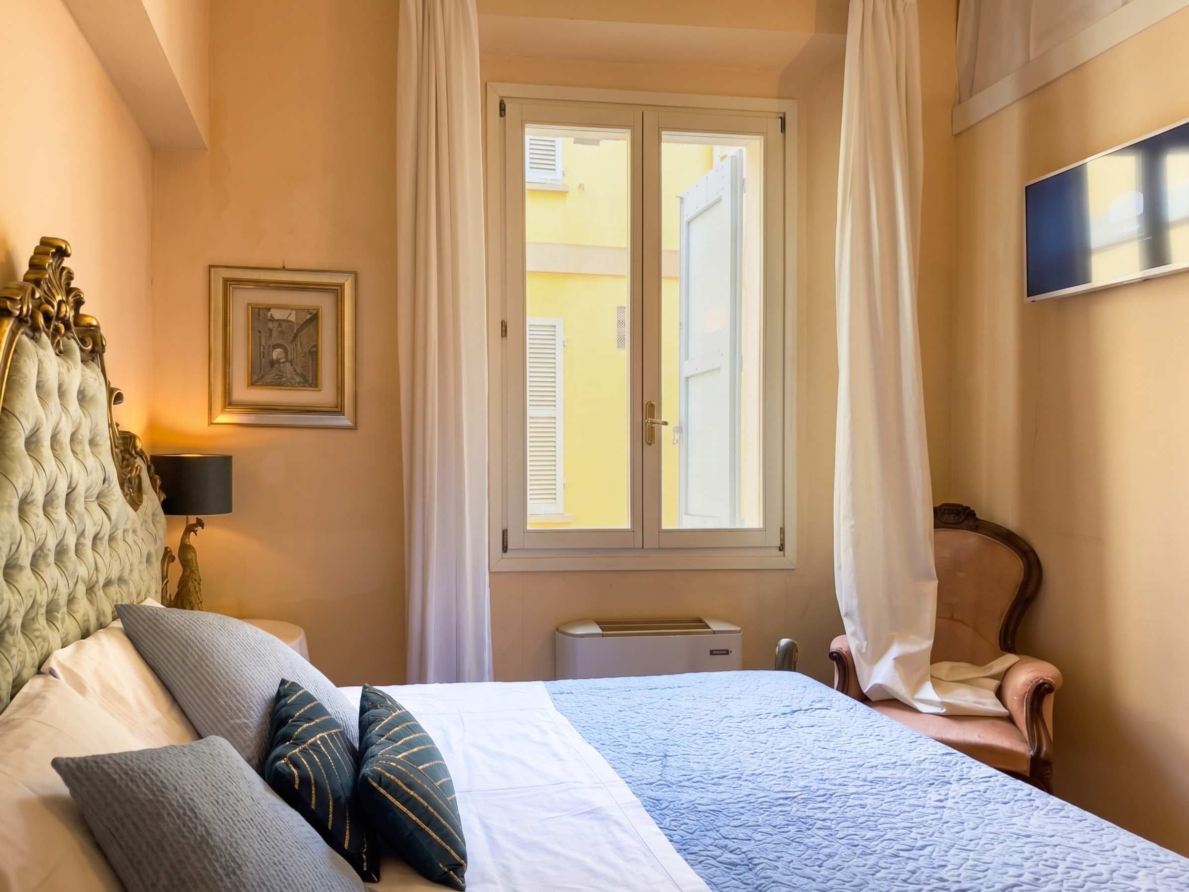 A hotel room with a bed, a chair, a window with curtains, a picture on the wall, and a flat-screen TV.