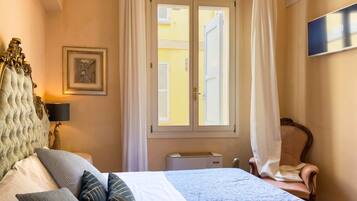 A hotel room with a bed, a chair, a window with curtains, a picture on the wall, and a flat-screen TV.