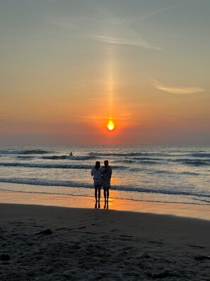 Cena de praia com duas pessoas em frente ao pôr do sol.