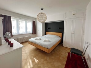 Quarto com cama de madeira, paredes brancas, radiador, escrivaninha com um ventilador e uma cadeira vermelha.