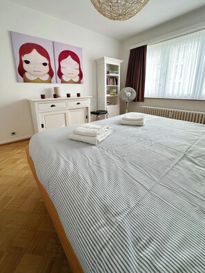 A neatly made bed with a striped bedspread, a white bedside table with a lamp, a white wardrobe, a bookshelf, and a window with curtains. - Sealia: a stunning flat on the seafront in Ostend with panoramic sea views (Oostende)