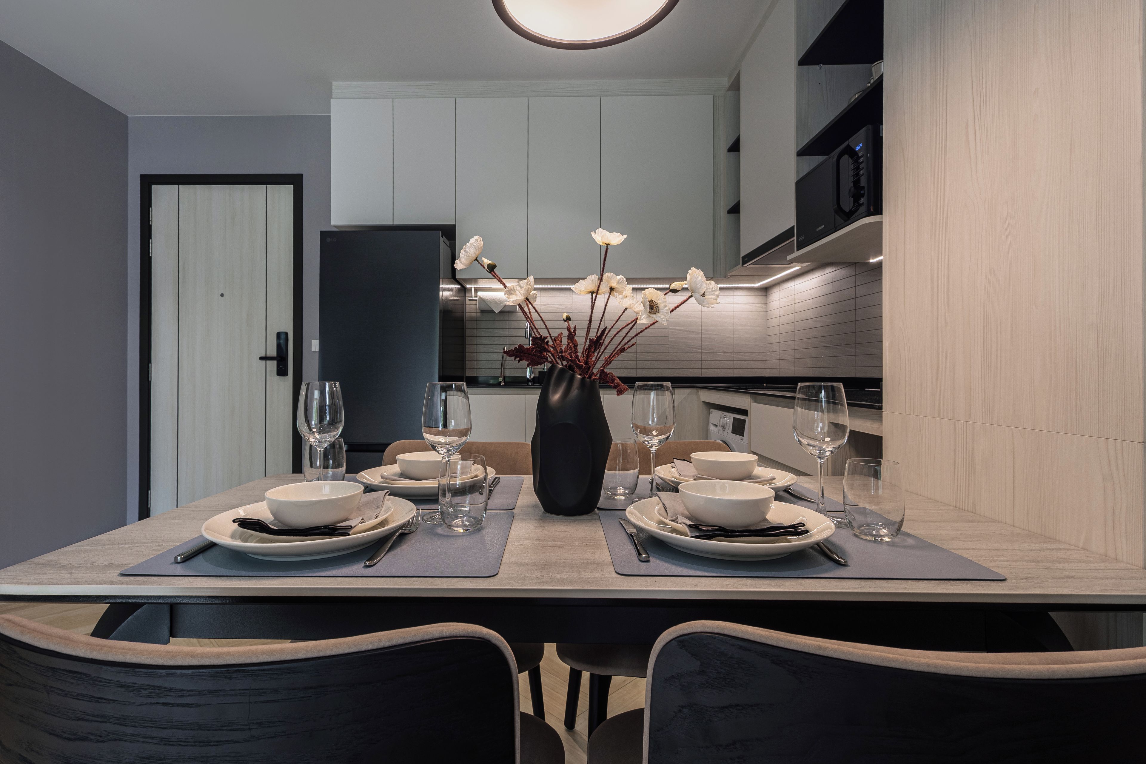 A modern dining area with a table set for two, a vase with flowers, and a kitchen in the background.