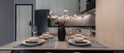 A modern dining area with a table set for two, a vase with flowers, and a kitchen in the background.