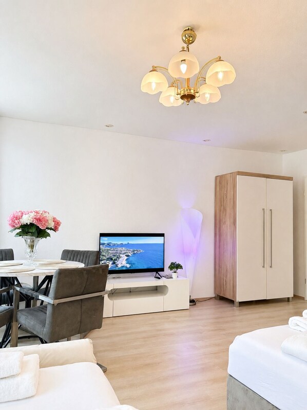 A modern living room with a dining area, a flat-screen TV, a chandelier, and a wardrobe.