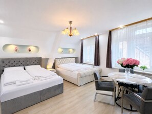A hotel room with two beds, a dining table with chairs, a chandelier, and a window with curtains.