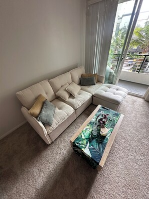 A living room with a beige sectional sofa, a coffee table with a tropical print, and a view of a balcony with greenery. - Luxe & Modern 1-bed with panel living space w Pool, Hot tub, Gym -Marina del Rey (Marina del Rey)