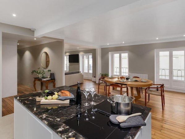 A modern kitchen with a marble countertop, a dining area with a round table, and a view of the outside through large windows.