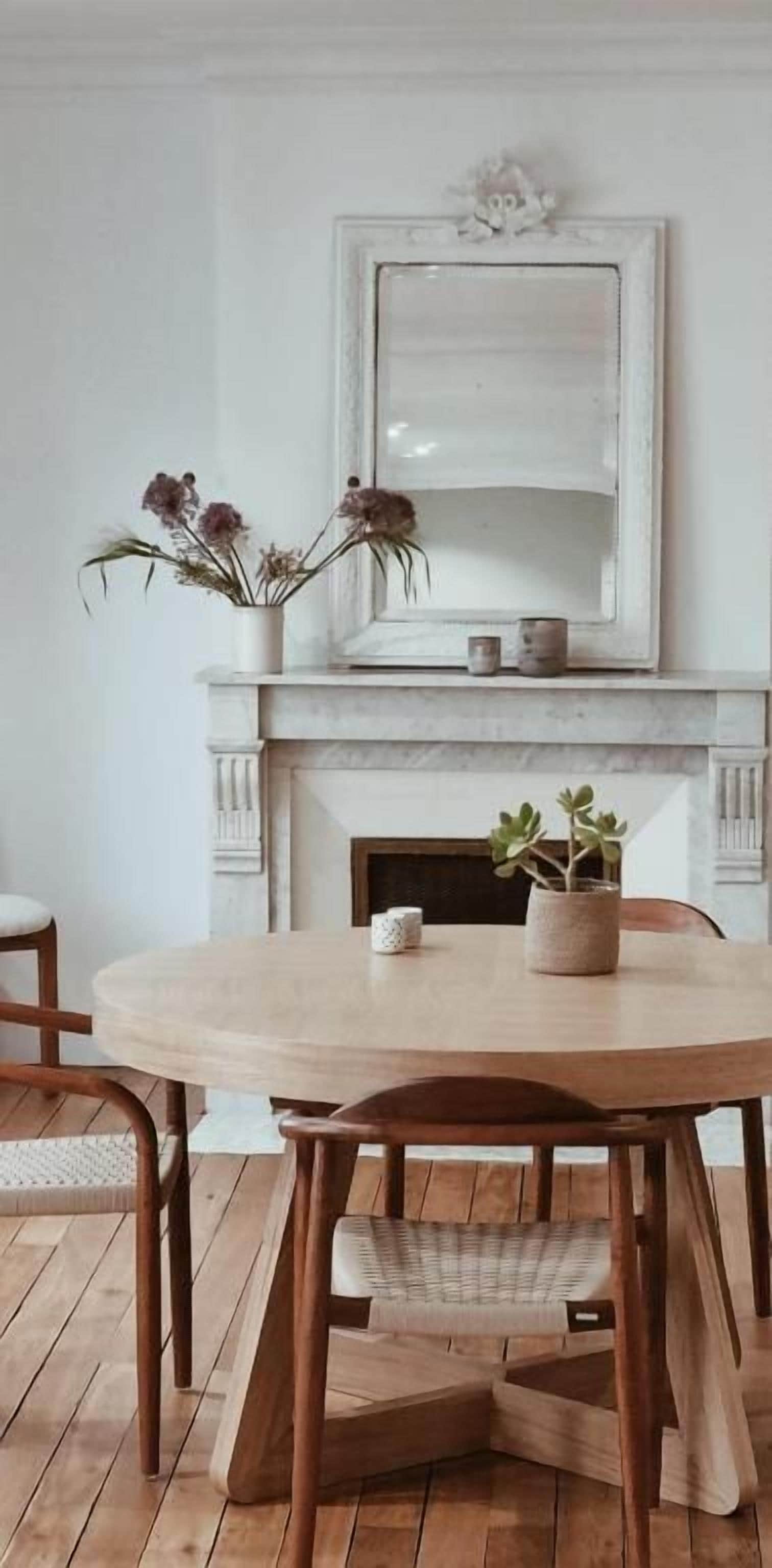 A dining area with a round wooden table, two chairs, a white vase with flowers, and a fireplace with a mirror above it.