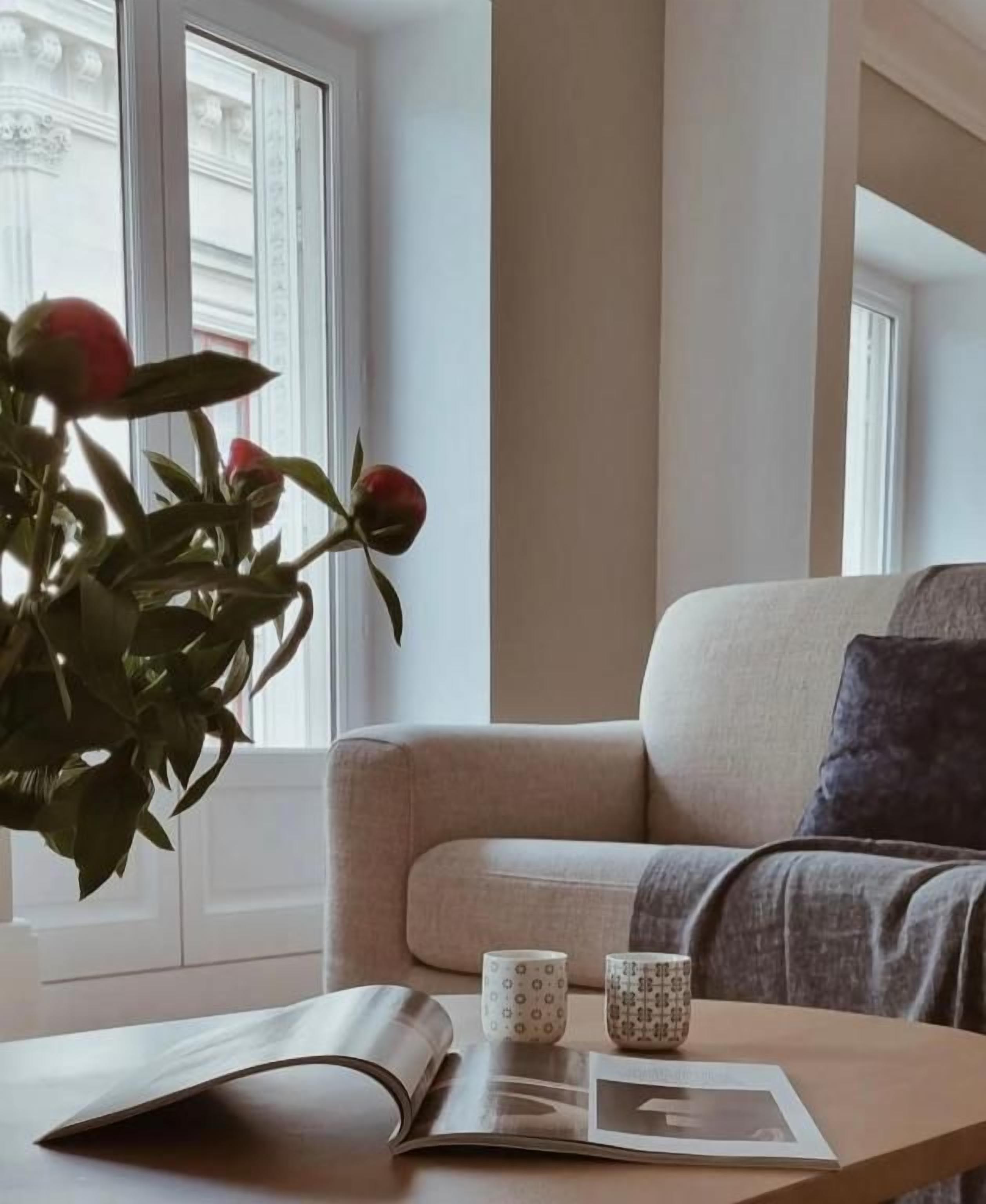 A room with a sofa, a table with a book and two mugs, and a window with a plant.