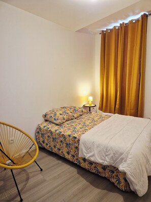 Ein Schlafzimmer mit einem Bett, einem Sessel, einem Nachttisch mit Lampe und Vorhängen.