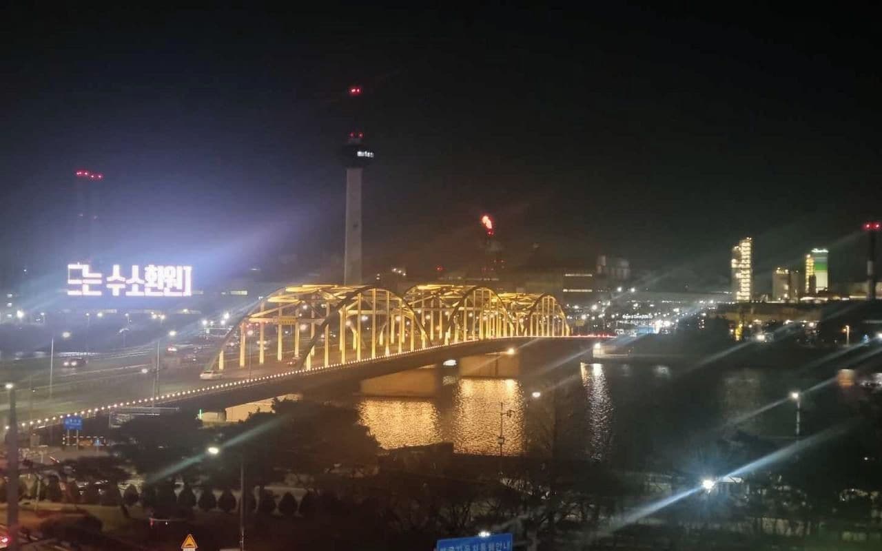 Vista nocturna de un puente con barandales iluminados y un cartel encendido al fondo.