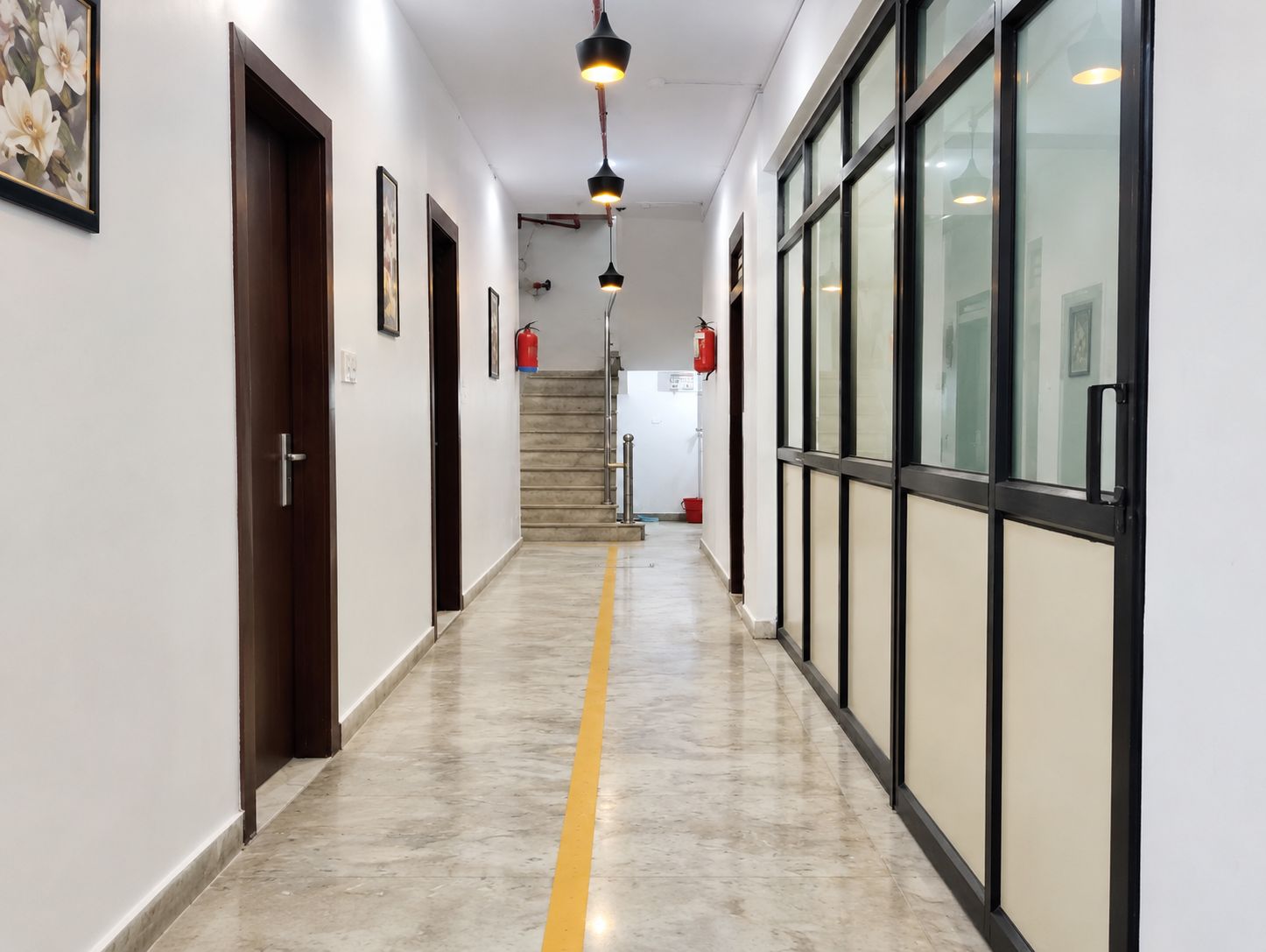 A hallway with a yellow line on the floor, doors, framed pictures, and fire extinguishers.