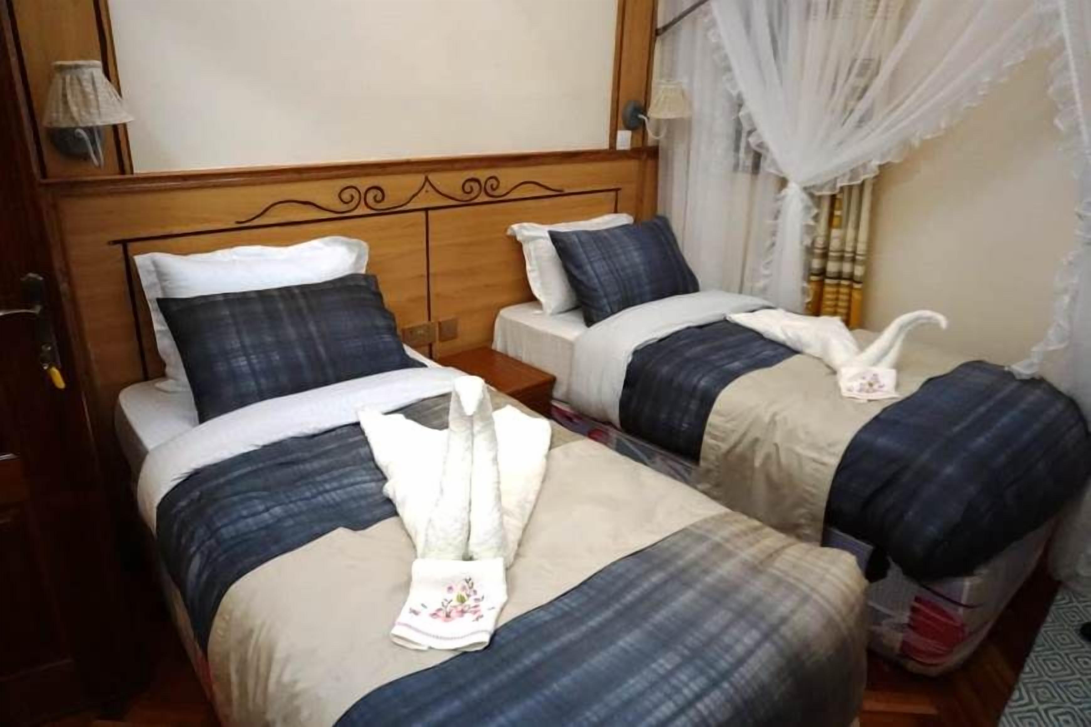 Una habitación de hotel con dos camas, cada una con cabecera, almohadas y manta. Hay una mesita de noche con una lámpara y una maleta.