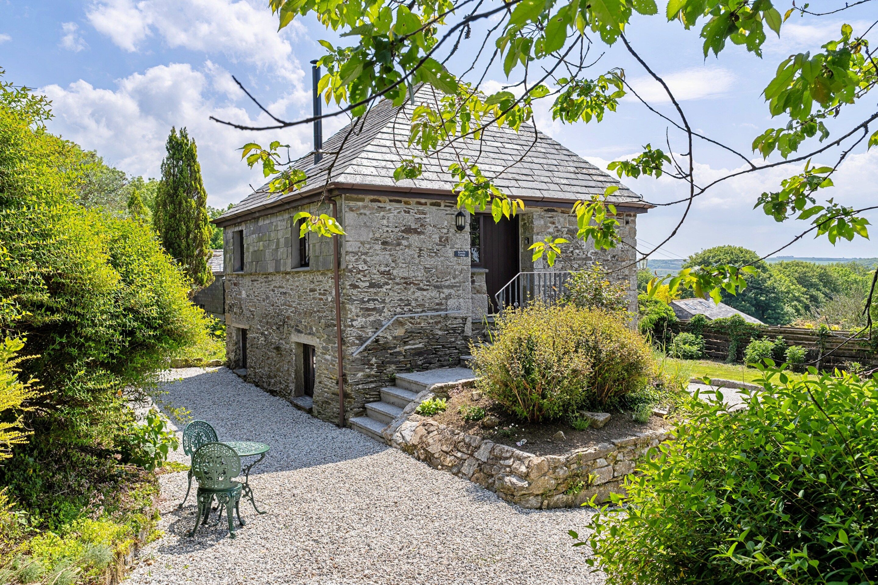 A stone house with a gravel driveway, a small garden, and a metal table and chair set.