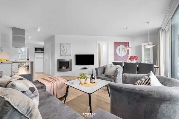 A modern living room with a grey sofa, a small coffee table, and a TV. There is a dining area in the background with a table and chairs. - Spacious holiday apartment with luxury features and covered terrace near the Bal (Lübeck)