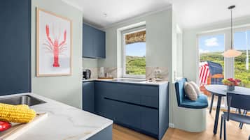 A modern kitchen with a built-in island, a dining area with a blue chair, and a large window offering a view of greenery.