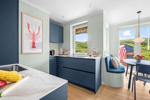Une cuisine moderne dotée d’un îlot central intégré, d’un coin repas avec une chaise bleue et d’une grande fenêtre offrant une vue sur la verdure.