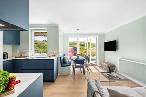 Une cuisine moderne dotée d’un four encastré, un coin repas avec une table et des chaises, et un espace de vie comprenant un canapé et un téléviseur fixé au mur.
