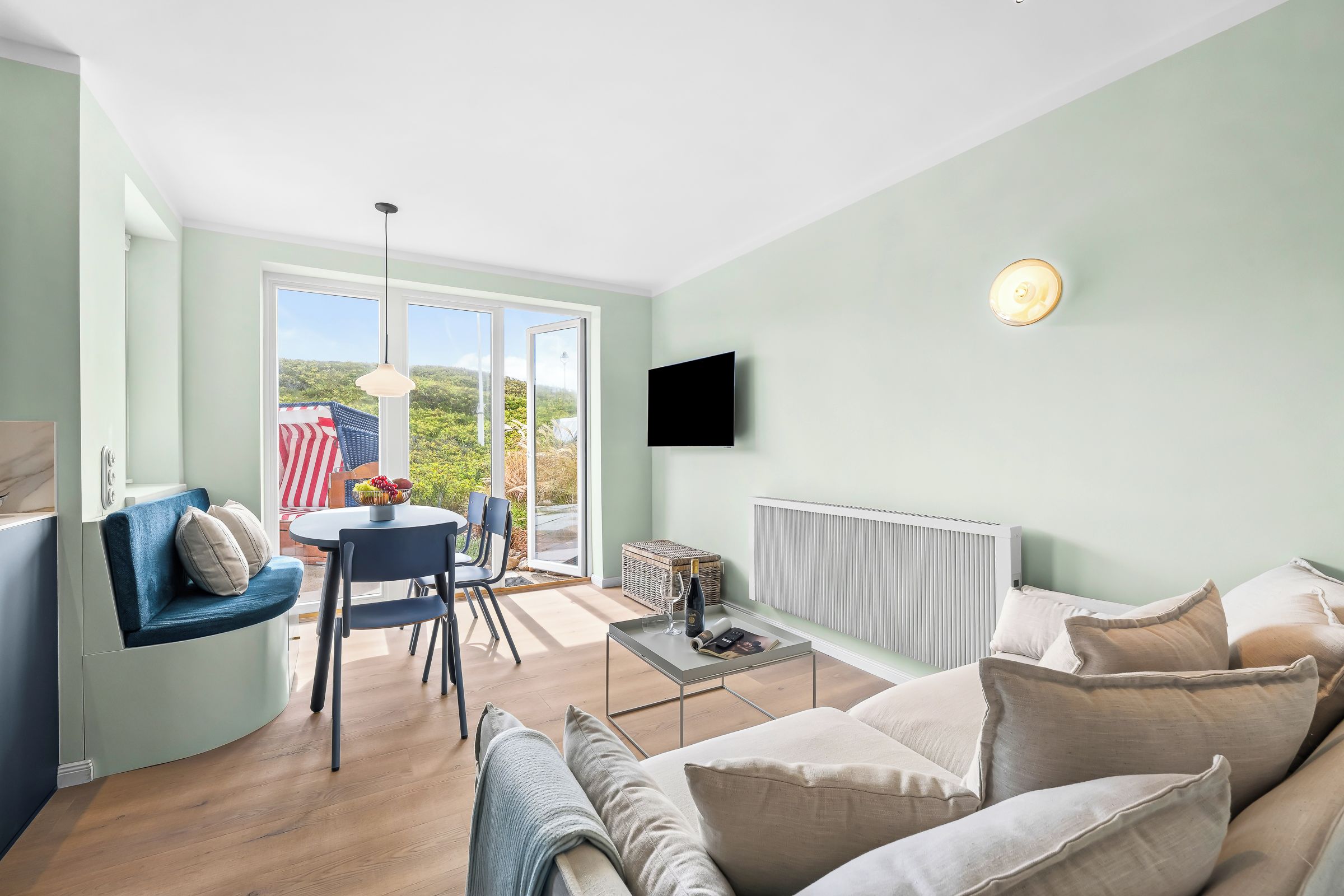 Una sala moderna con sofá, una mesa de comedor y sillas. Hay un televisor montado en la pared y una ventana con vistas a zonas verdes.