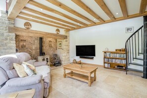 A cozy living room with a stone wall, a fireplace, a sofa, a coffee table, a bookshelf, and a TV. - Holly Cottage - Beautiful 3 Bedroom Cottage near Bristol (Backwell)