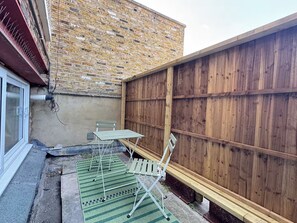 Eine Terrasse mit Ziegelwand, Holzzaun und Gartenmöbeln, darunter ein Tisch und Stühle.