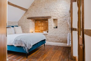 A bedroom with a stone wall, wooden beams, a bed with a blue cover, and a small window.