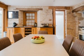 Un coin repas avec une table dressée pour quatre personnes, un panier de fruits sur la table, et une cuisine aux murs en pierre et aux armoires en bois.