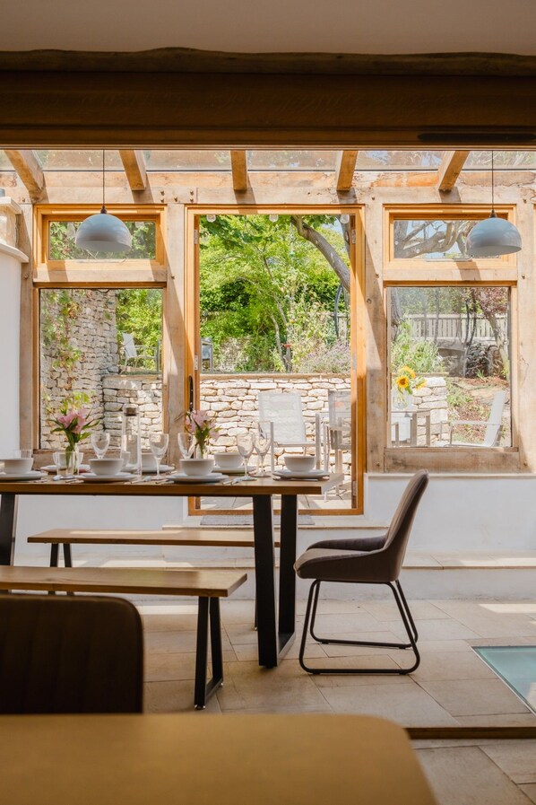 A dining area with a table set for two, wooden beams, and a view of a stone wall and greenery outside.