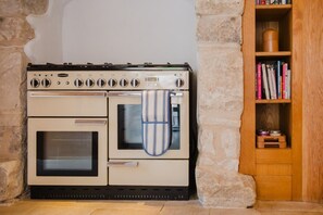 A kitchen with a double oven, a kettle, and a wooden bookshelf with books.
