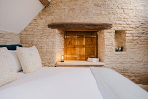 Un dormitori amb paret de pedra, porta de fusta, llit amb roba de llit blanca i una petita estant amb tovalloles.
