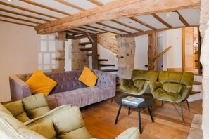 Une pièce avec un plafond en bois, un canapé avec des coussins, deux fauteuils et un escalier en arrière-plan.