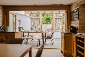 Une cuisine avec des meubles en bois, une table à manger dressée pour quatre personnes et une vue sur le jardin par des portes ouvertes.