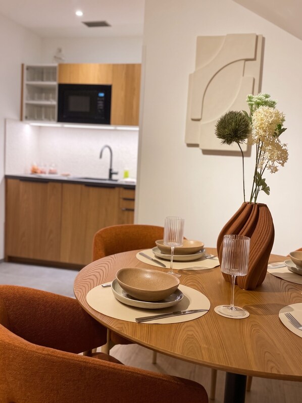 A modern dining area with a wooden table set for two, a vase with flowers, and a kitchen in the background.