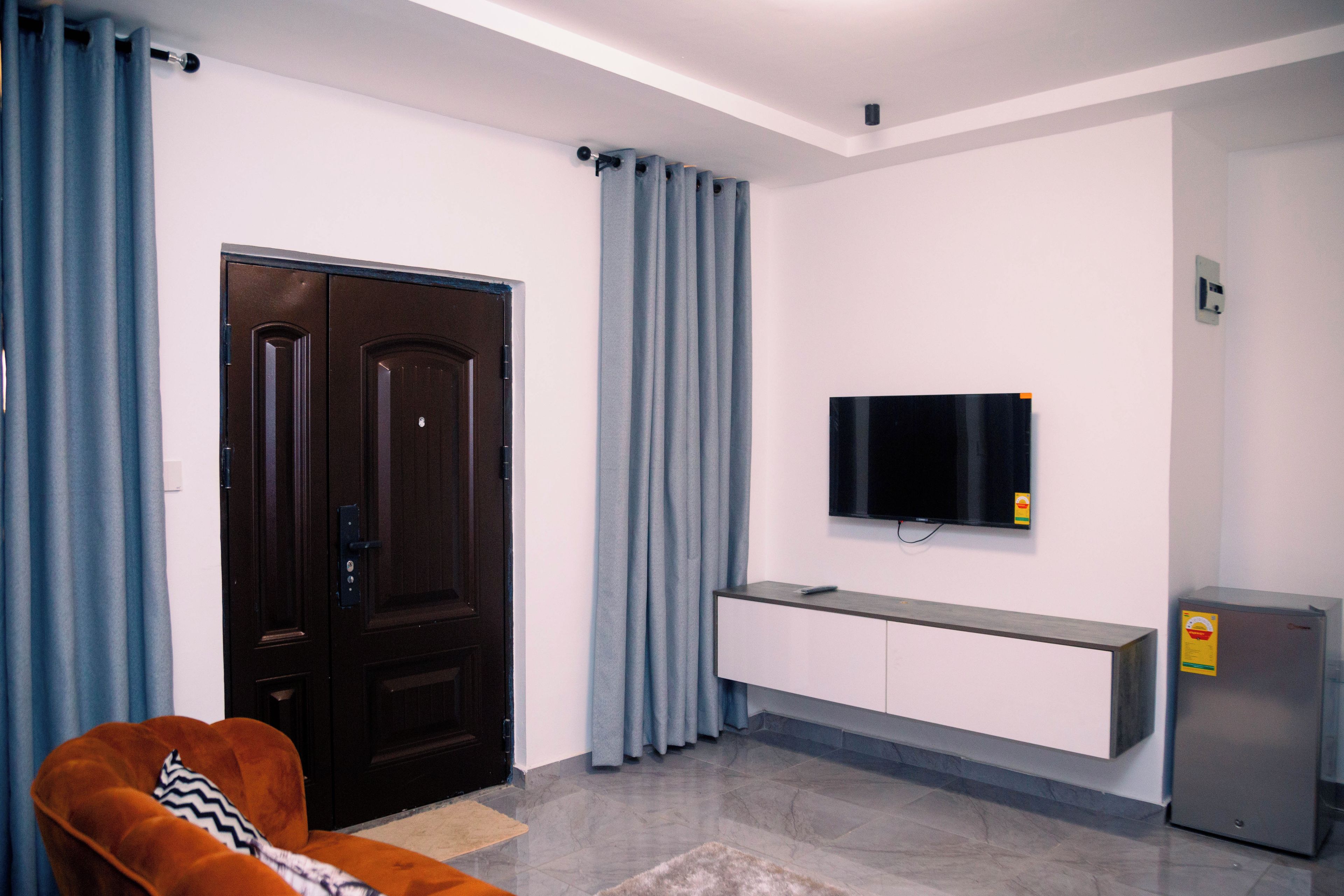 A modern living room with a dark wooden door, light blue curtains, a flat-screen TV mounted on a white cabinet, and a brown sofa.